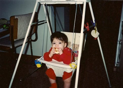A baby in swinging chair stares directly into the camera while munching on a mini-bagel.