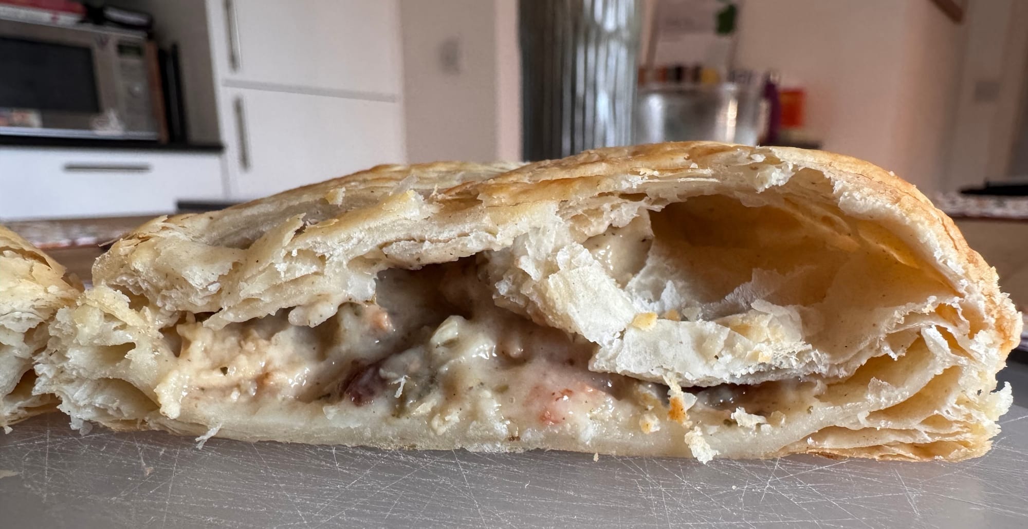 A Vegan Festive Bake cut in half on a cutting board. Puffy pastry layers and the creamy festive internals are clearly on display. The filling is very liquidy, though it is not spilling out in an unmanagable way.