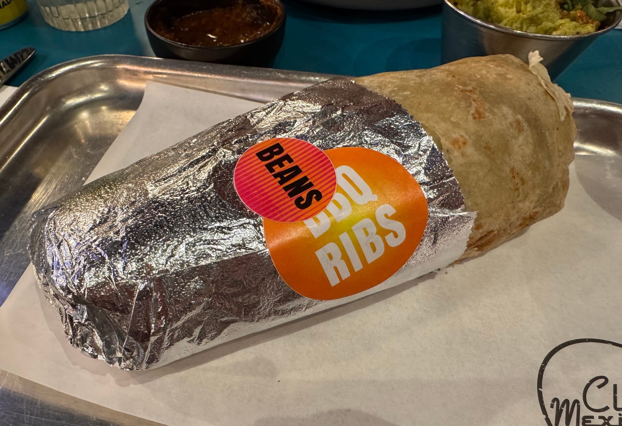 The Club Mexicana BBQ ribs burrito, presented like street food, wrapped in tin foil with two stickers reading "BBQ Ribs" and "Beans."