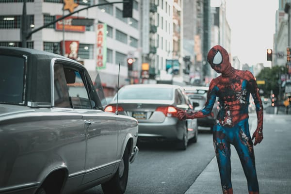 A person in a beat-up Spider-Man costume thumbs for a ride on the sidewalk in a city.